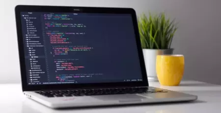 A laptop displays code in a text editor, showcasing a JavaScript file related to user registration. In the background, there is a small potted plant and a yellow mug on a white desk.