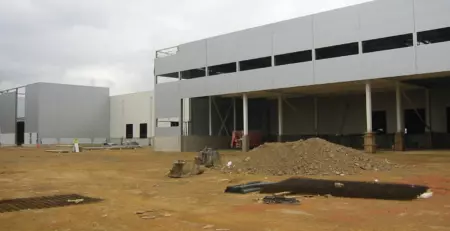 A construction site features partially completed buildings with gray exteriors and large windows. The ground is uneven with piles of dirt and construction materials scattered around.