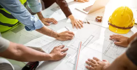 A group of professionals collaborates around a table covered with architectural plans and blueprints. Tools and safety equipment, including a yellow hard hat, are visible, indicating a construction or engineering project discussion.