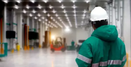 A worker in a green jacket and white hard hat stands in a spacious warehouse, observing the activity in the background. The well-lit environment features forklifts and other equipment, indicating a busy industrial setting.