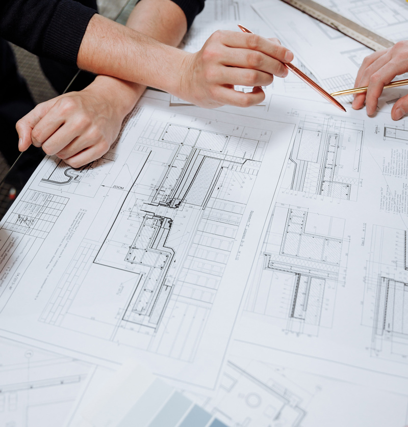 Two hands are seen discussing architectural plans laid out on a table. The plans feature detailed drawings and measurements, indicating a collaborative design process.