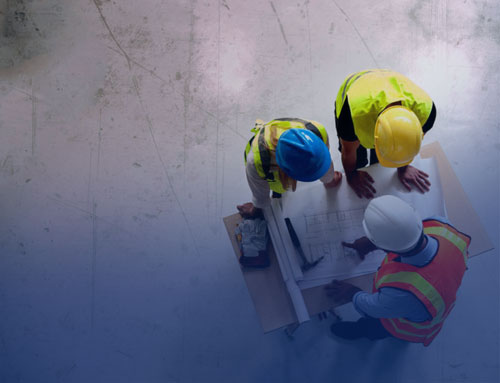 Three construction workers are gathered around a set of blueprints on a table. They are wearing safety helmets and vests, discussing plans for a project.