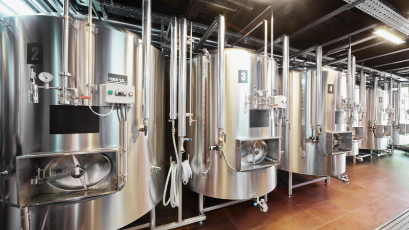 A row of large, shiny stainless steel fermentation tanks is displayed in a modern facility. Each tank is labeled with a number and features various valves and gauges for monitoring.