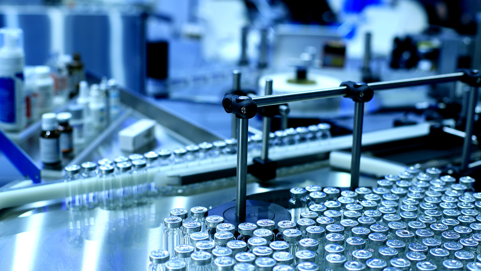 A collection of glass vials is arranged on a metallic surface, surrounded by various pharmaceutical products. The setting appears to be a laboratory or manufacturing facility focused on medication preparation.