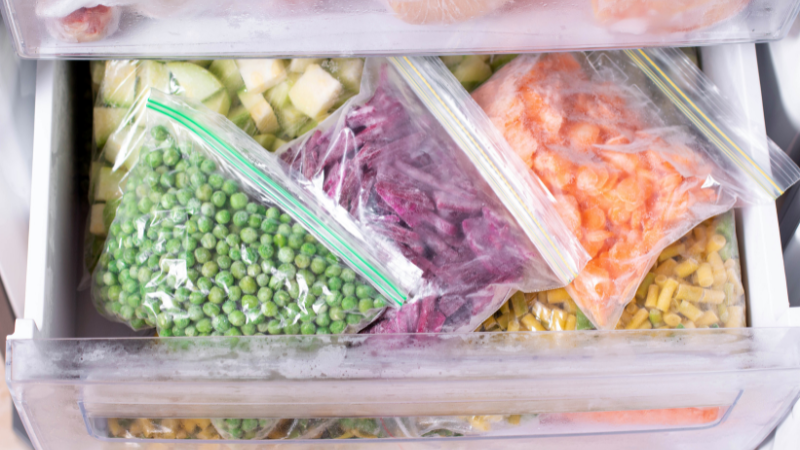 A freezer drawer filled with various bags of frozen vegetables and fruits, including green peas, diced carrots, and mixed fruit. The items are neatly organized in clear plastic bags, showcasing a variety of colors and textures.