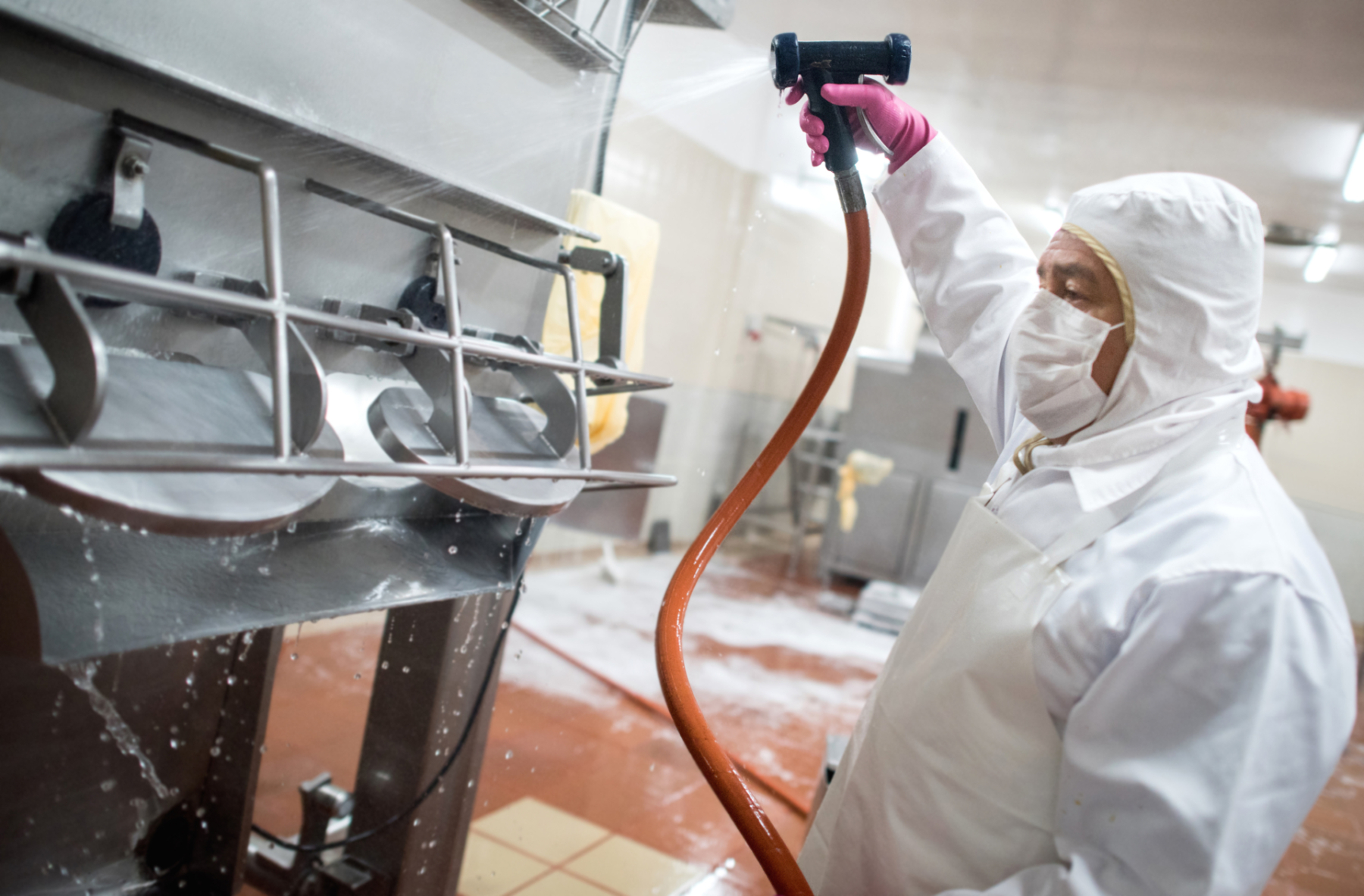 A worker in protective clothing is using a high-pressure hose to clean industrial equipment in a food processing facility. Water and soap are spraying from the equipment, highlighting the sanitation process.