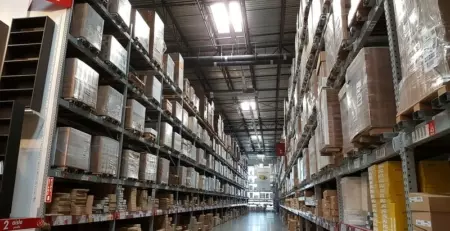 A wide aisle in a warehouse filled with tall shelves stacked with boxes and pallets. The ceiling is high, and bright lights illuminate the space, creating a spacious and organized environment.