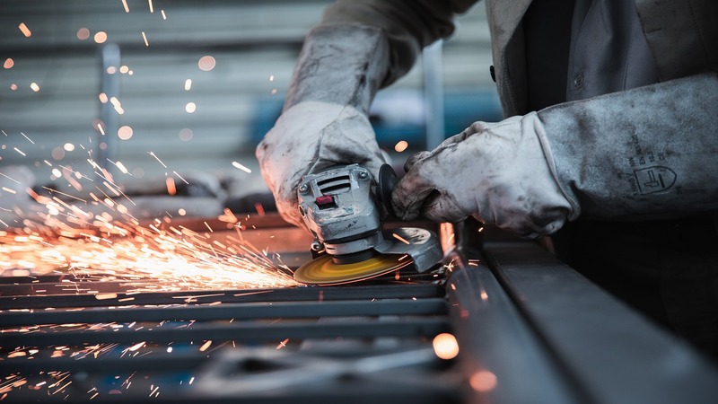 A person wearing protective gloves operates a power tool, creating sparks as they grind metal. The scene captures the intensity and focus of metalworking in a workshop environment.