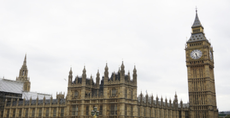 The image features the iconic architecture of the Palace of Westminster, with its intricate towers and detailed stonework. The prominent clock tower, known as Big Ben, stands tall against a cloudy sky.