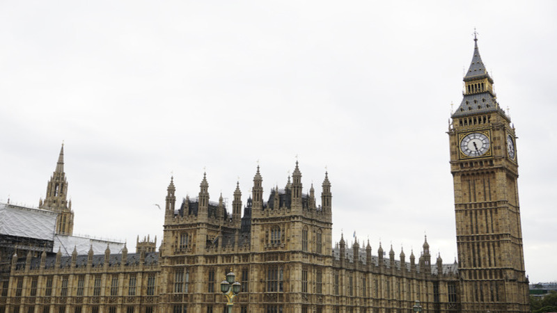 The image features the iconic architecture of the Palace of Westminster, with its intricate towers and detailed stonework. The prominent clock tower, known as Big Ben, stands tall against a cloudy sky.