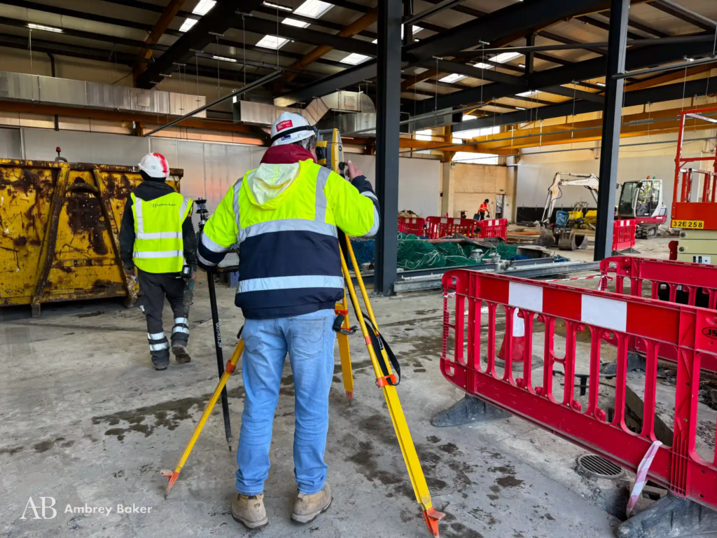 Two workers in safety gear are on a construction site, one using a surveying instrument while the other walks away. The background features construction equipment and safety barriers.