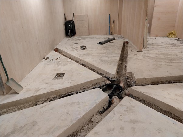 A construction site features a concrete floor with several trenches and a central drainage system. Tools and equipment are scattered around, indicating ongoing work in the space.