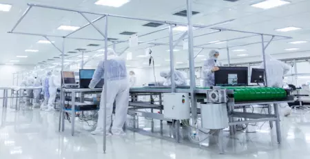 A cleanroom environment is depicted, with several individuals in protective suits working at tables equipped with computers and machinery. The bright, sterile space emphasizes safety and precision in a technical setting.