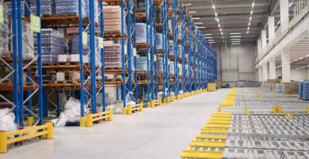 A spacious warehouse filled with tall shelving units stacked with various goods. The floor is clear, with safety barriers marking the pathways between the shelves.