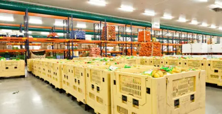 A spacious warehouse filled with yellow crates containing various fruits and vegetables. Shelves in the background are stocked with colorful produce and supplies.