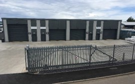 A row of storage units with gray metal doors is visible, set against a cloudy sky. A gated entrance leads to the concrete area in front of the units.