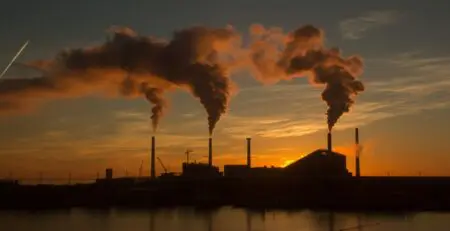 Silhouetted against a vibrant sunset, industrial smokestacks release thick plumes of smoke into the sky. The scene captures the contrast between nature and industrial activity, with a calm body of water reflecting the colors of the sunset.