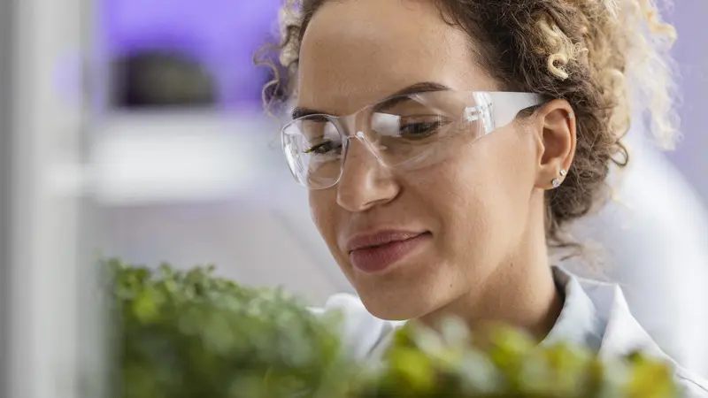 A woman wearing safety glasses examines green plants with a focused expression. She appears to be engaged in scientific research or cultivation.