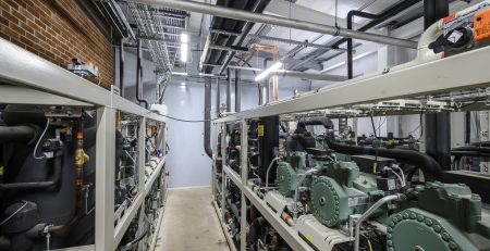 The image features a mechanical room filled with large industrial equipment, including multiple compressors and piping systems. The space is well-lit, with visible electrical conduits and a brick wall in the background.