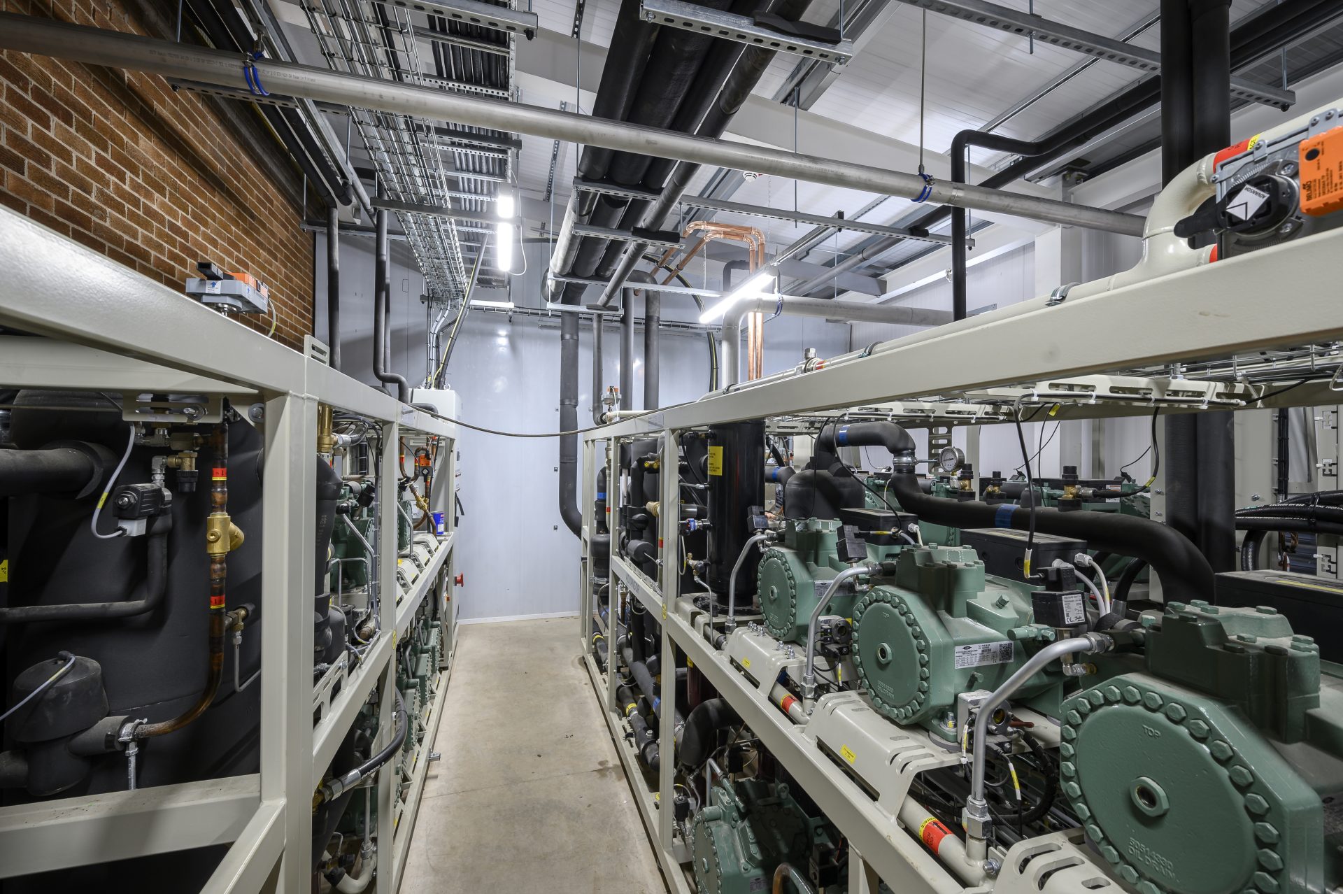 The image features a mechanical room filled with large industrial equipment, including multiple compressors and piping systems. The space is well-lit, with visible electrical conduits and a brick wall in the background.