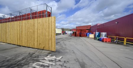 A wide view of a storage area featuring wooden fencing and stacked crates in various colors. The scene is set under a partly cloudy sky, with a clear pathway leading into the space.