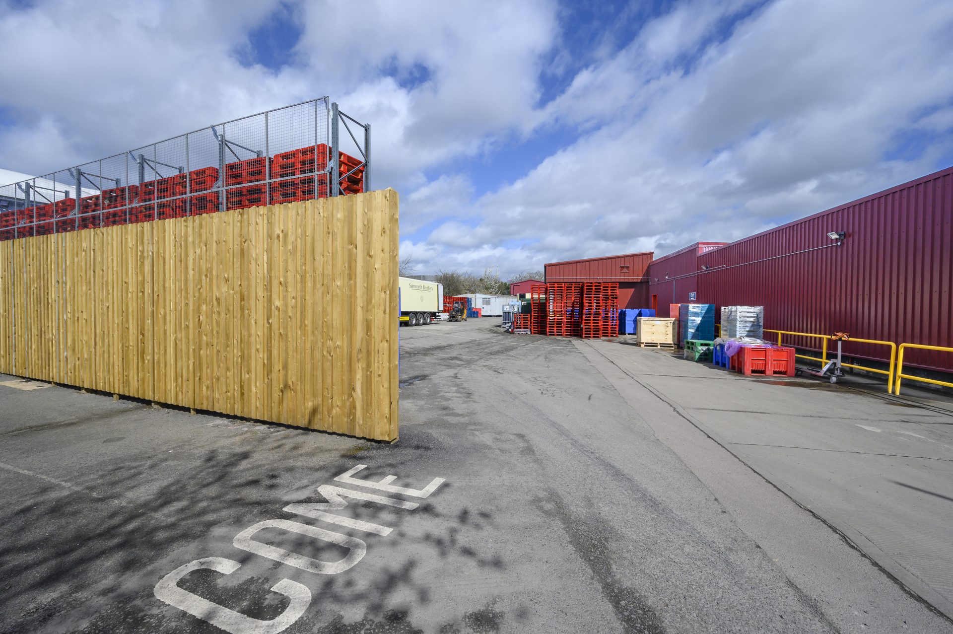 A wide view of a storage area featuring wooden fencing and stacked crates in various colors. The scene is set under a partly cloudy sky, with a clear pathway leading into the space.