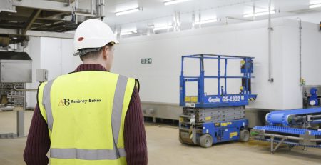 A person wearing a safety helmet and a high-visibility vest stands in a spacious industrial area. Equipment such as a scissor lift and conveyor system is visible in the background.