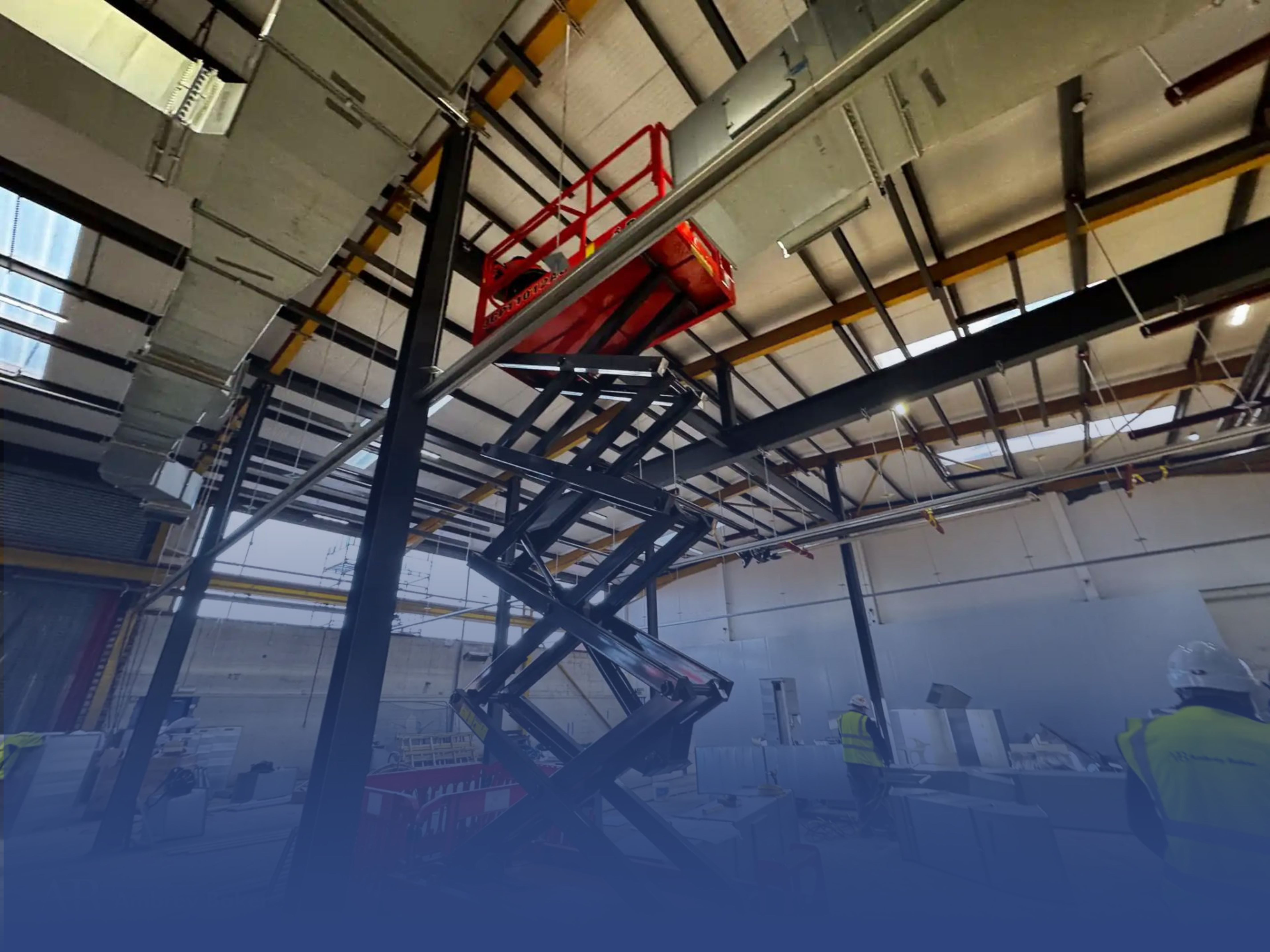 A scissor lift is positioned high in a warehouse, with workers in safety gear visible below. The interior features exposed beams and ductwork, indicating ongoing construction or maintenance.