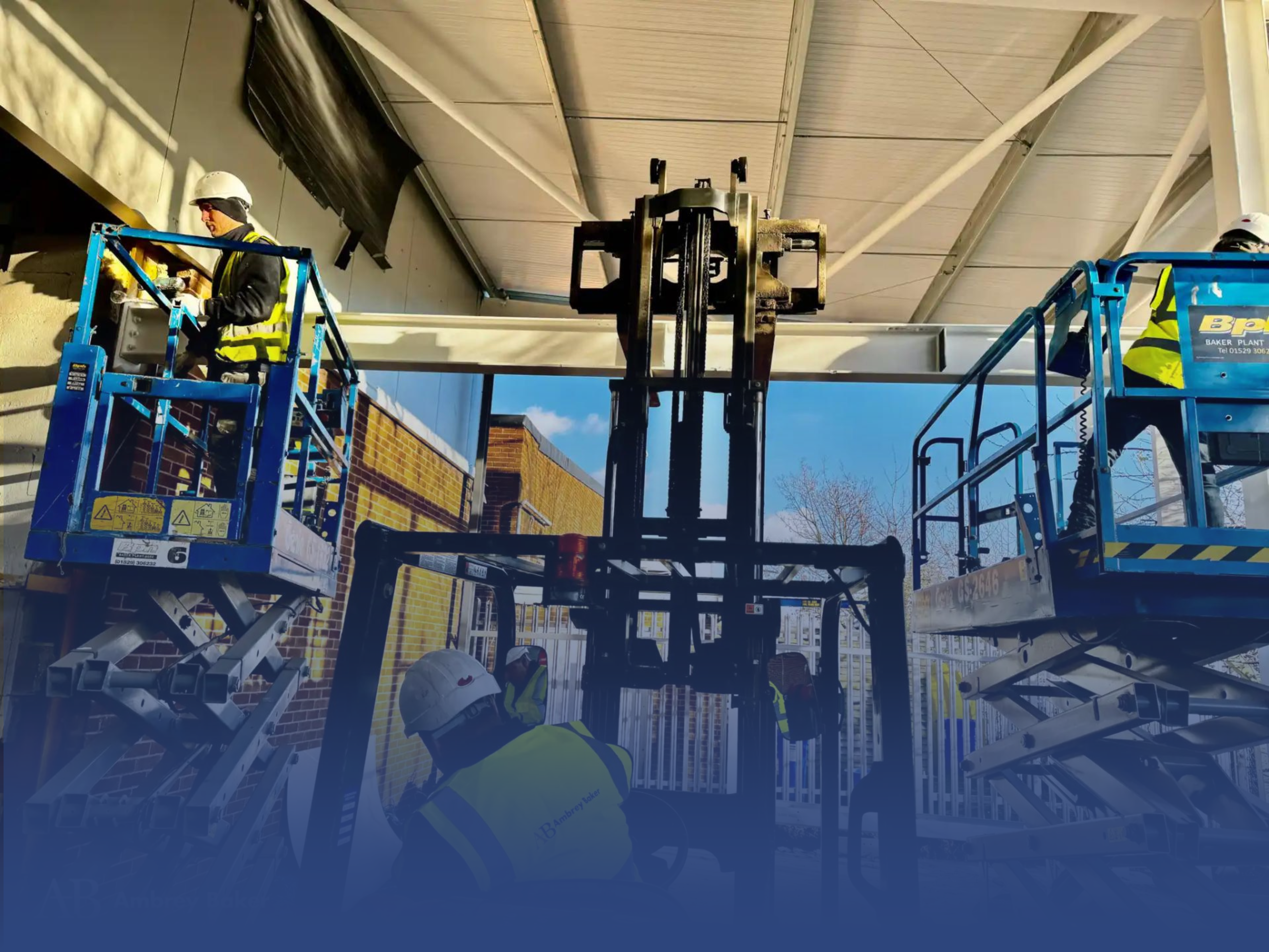 Workers are using scissor lifts and a forklift to perform construction tasks at a building site. The scene shows a clear blue sky and a partially completed structure.