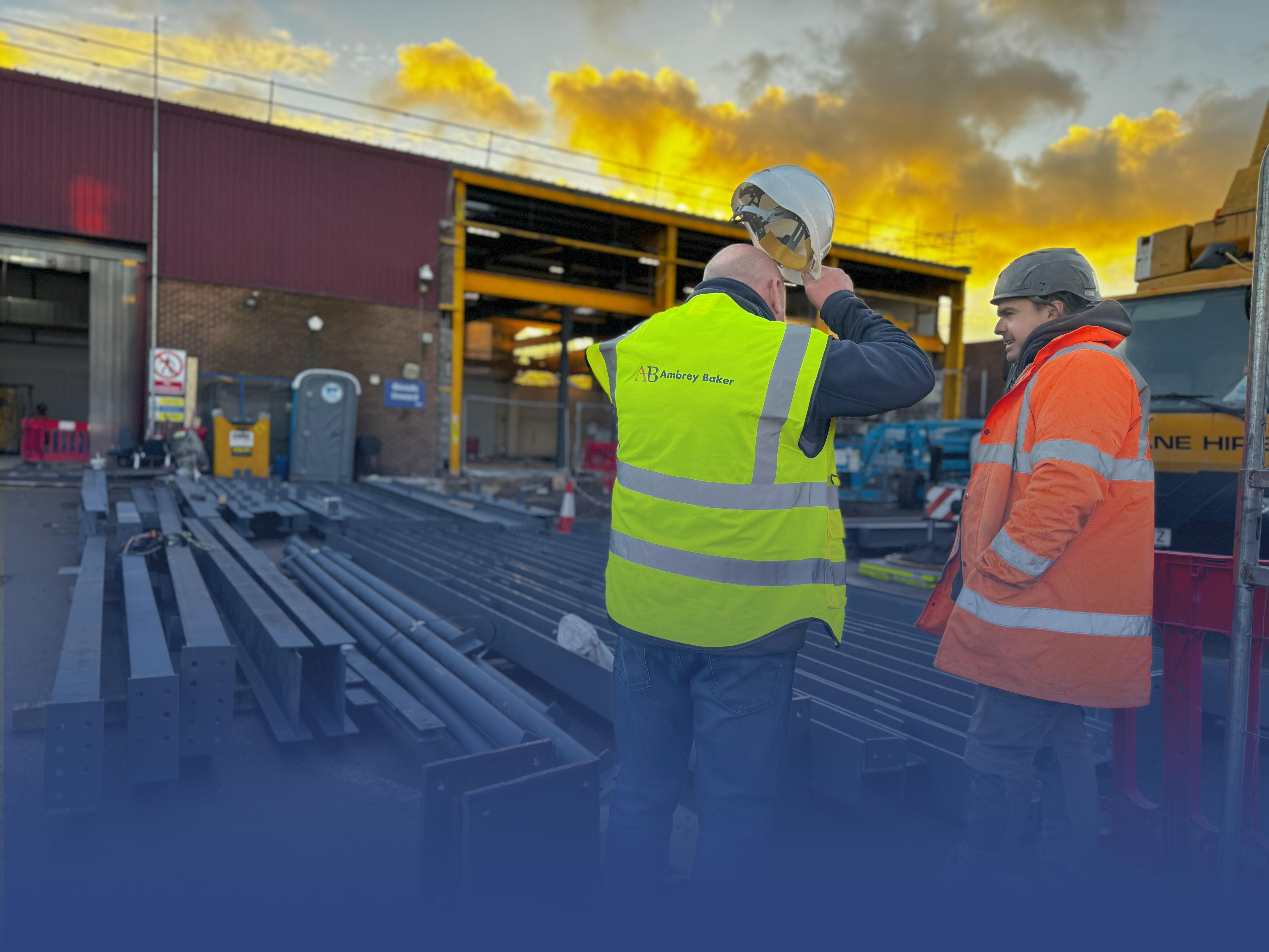 Two construction workers are engaged in conversation on a site, surrounded by steel beams and machinery. The sky in the background features vibrant clouds, indicating either sunrise or sunset.