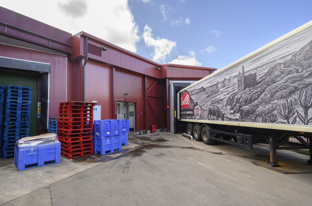 A delivery truck is parked at a loading dock, with colorful pallets stacked nearby. The building features a red exterior and a mural depicting a coastal scene.