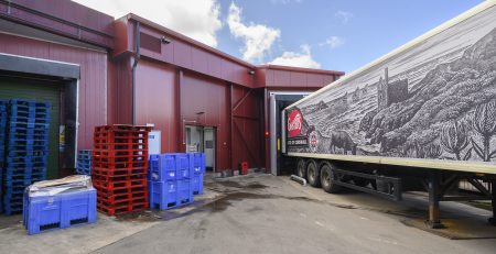 A delivery truck is parked at a loading dock, with colorful pallets stacked nearby. The building features a red exterior and a mural depicting a coastal scene.