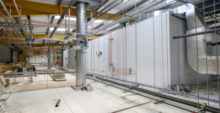 An industrial space under construction features exposed ductwork, pipes, and electrical installations. The floor is partially finished, with various materials and tools scattered throughout the area.