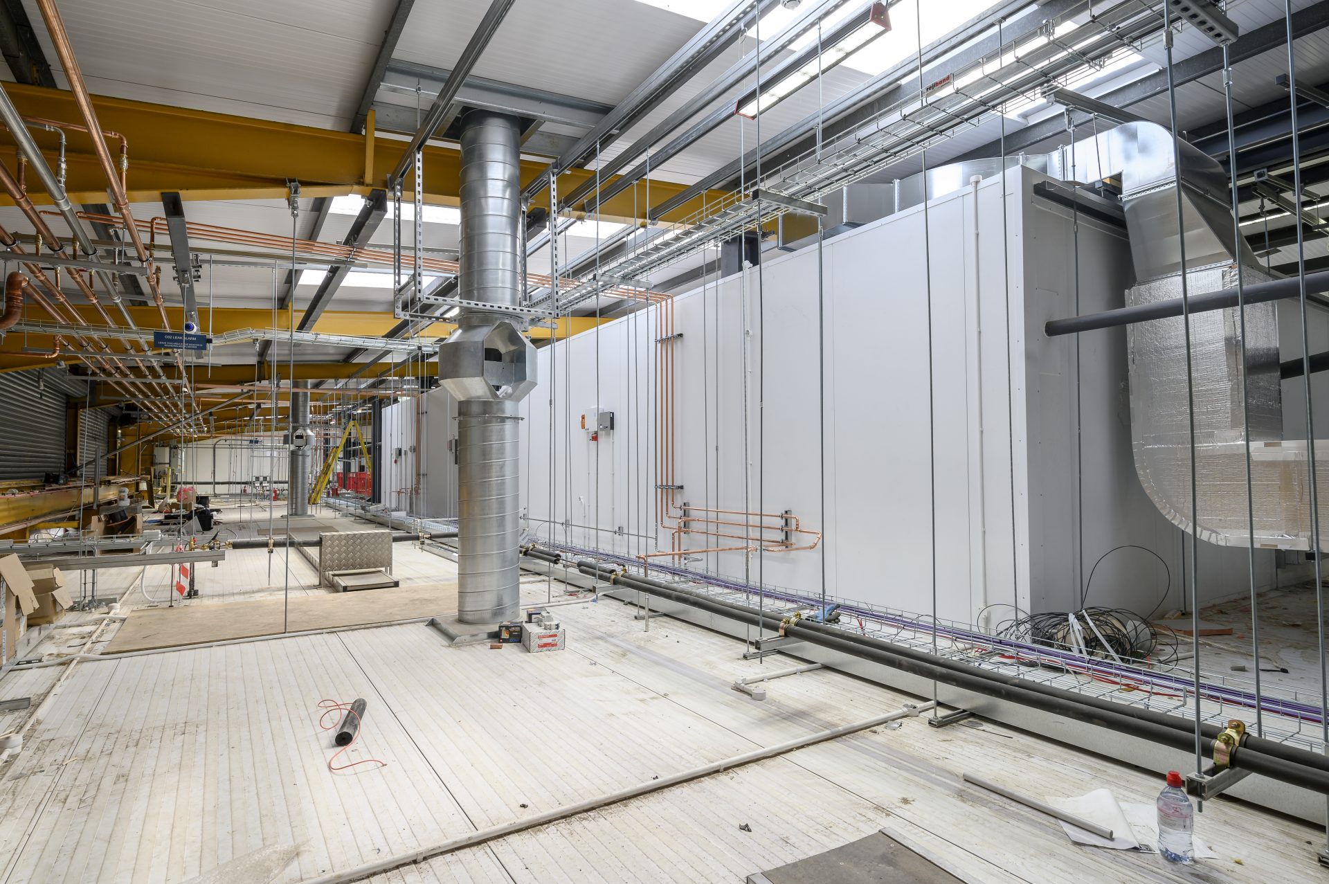 An industrial space under construction features exposed ductwork, pipes, and electrical installations. The floor is partially finished, with various materials and tools scattered throughout the area.