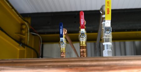 Three valves in different colors—blue, red, and yellow—are mounted on copper piping. The valves are part of a plumbing system, with visible connections and a metallic structure in the background.