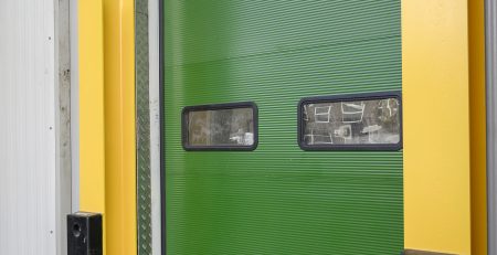 A bright green industrial door is framed by yellow side panels and features two small windows. The door is partially open, revealing a glimpse of the interior space.