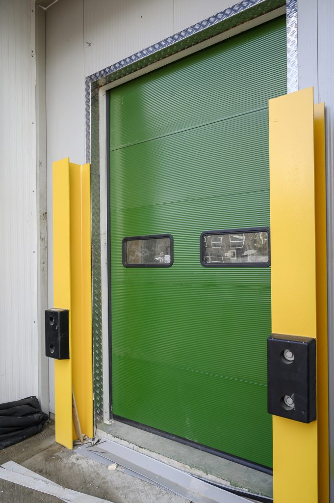 A bright green industrial door is framed by yellow side panels and features two small windows. The door is partially open, revealing a glimpse of the interior space.