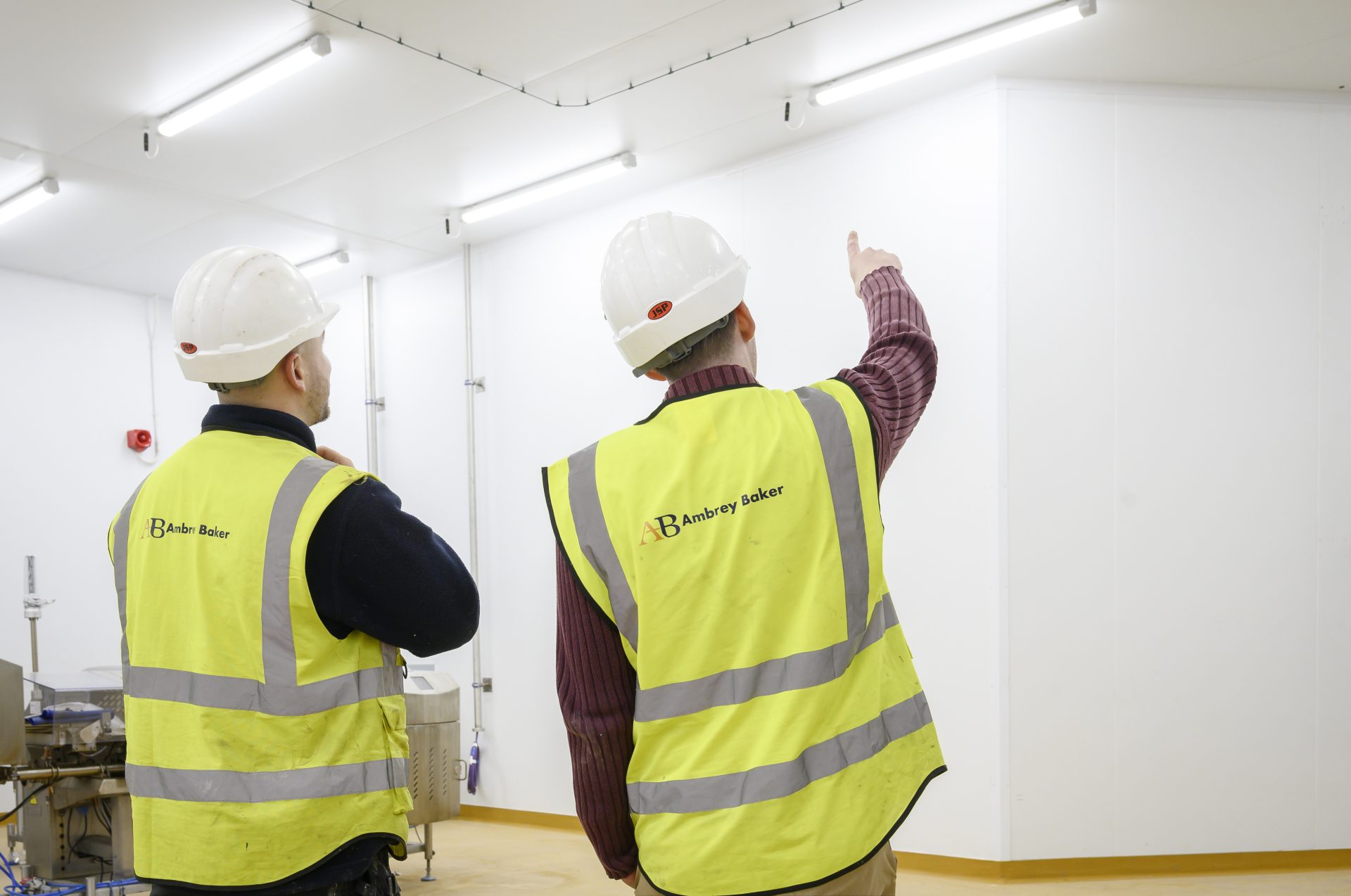 Two individuals in safety vests and hard hats are discussing something in a well-lit, industrial space. One person is pointing towards the ceiling, indicating an area of interest.